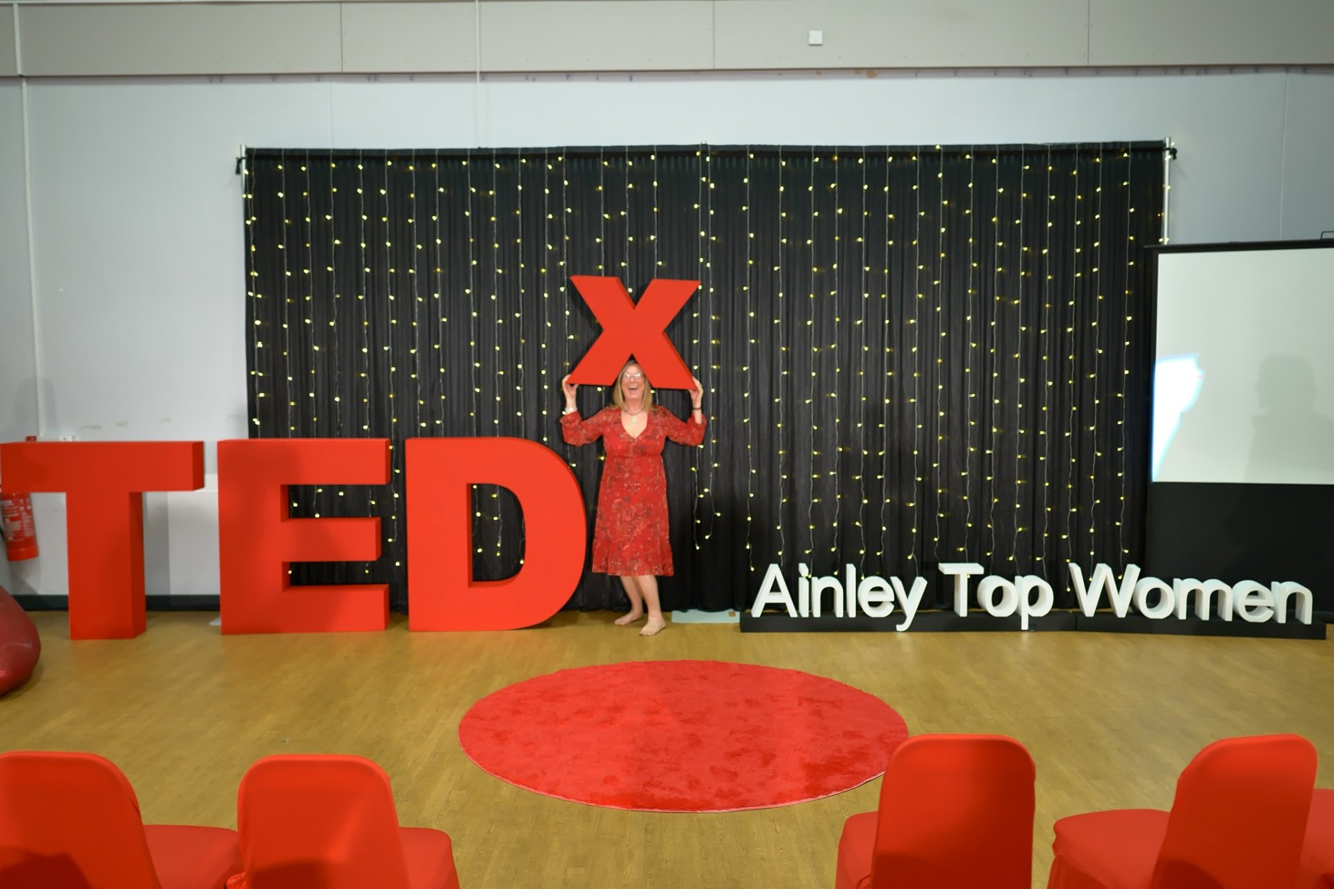 Marion Bevington on the TEDx AinleyTopWomen stage in a red dress, holding the giant red X letter above her head, laughing.